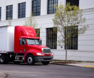 Red semi truck with short day cab and high spoiler to improve aerodynamic of rig with dry van trailer delivering goods by urban city street with contemporary buildings.